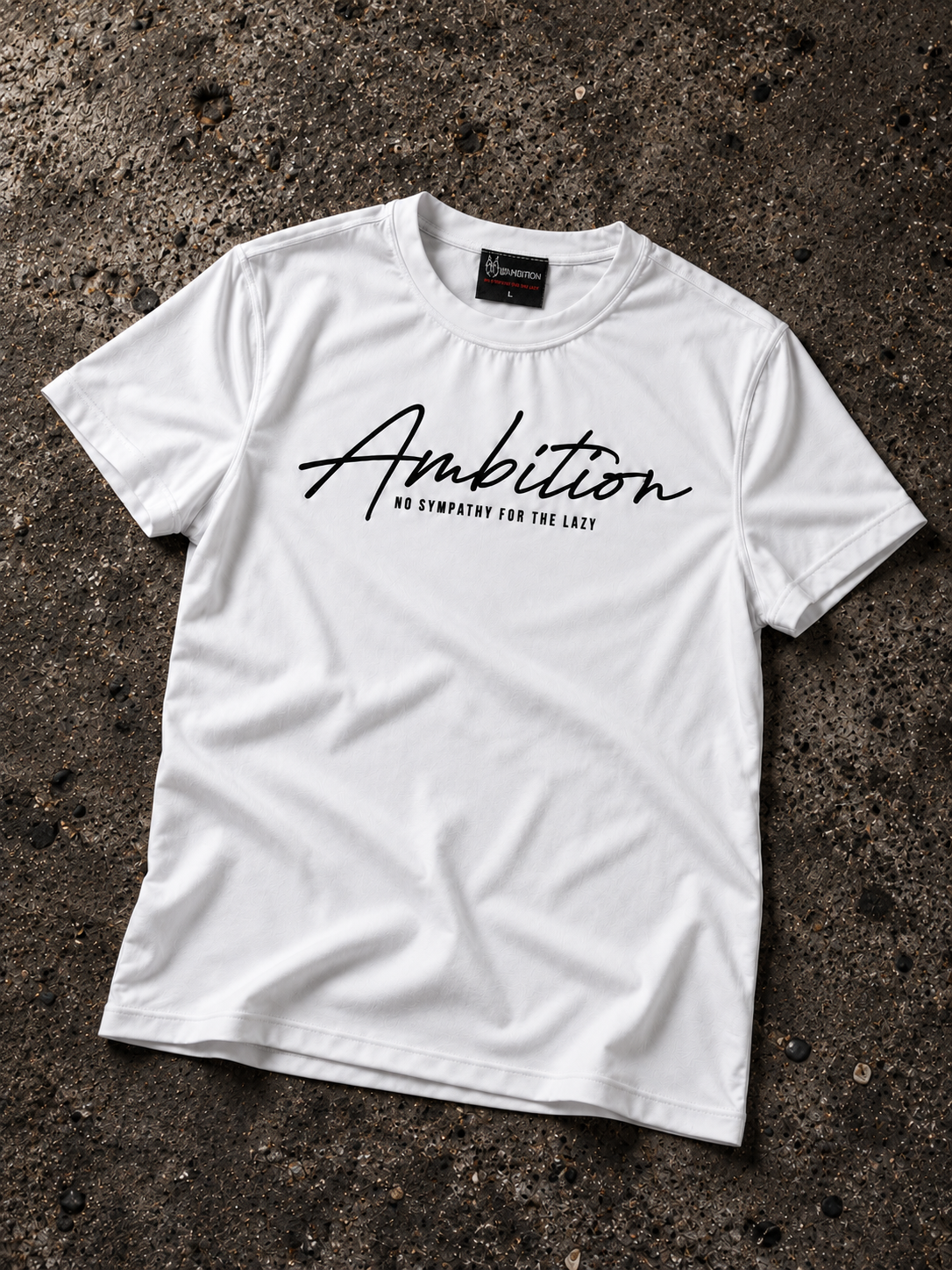 White Ambition T-shirt laid flat on dark textured concrete, showing the black chest print, small tagline and sewn neck label.